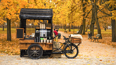 Cargo bike adibita a carretto del caffè parcheggiata in un parco autunnale, con foglie arancioni e gialle a terra e atmosfera accogliente